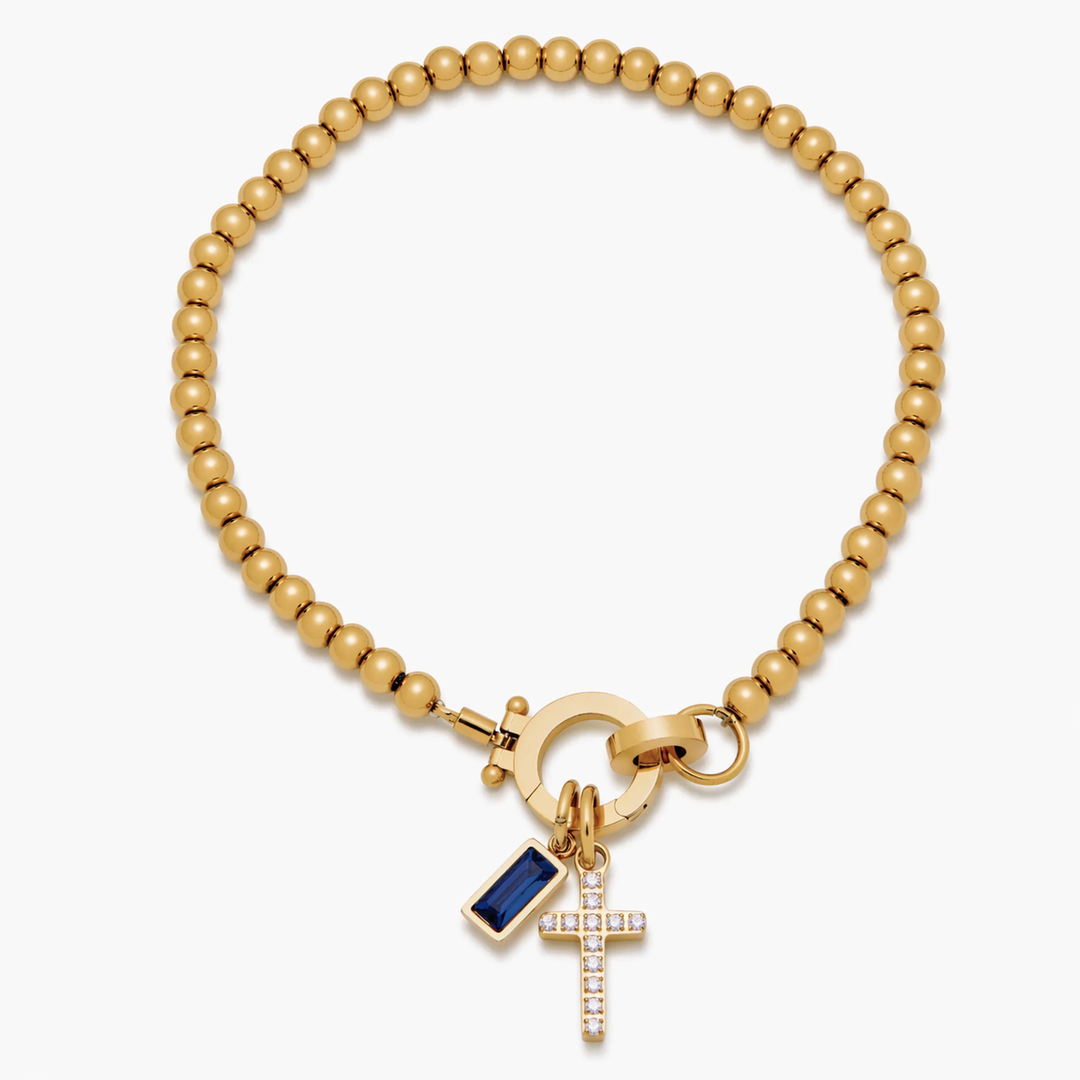 Pulsera de Auxana con dije de cruz y piedra natal, hecha de metal y piedra colorida, con diseño elegante y simbólico.